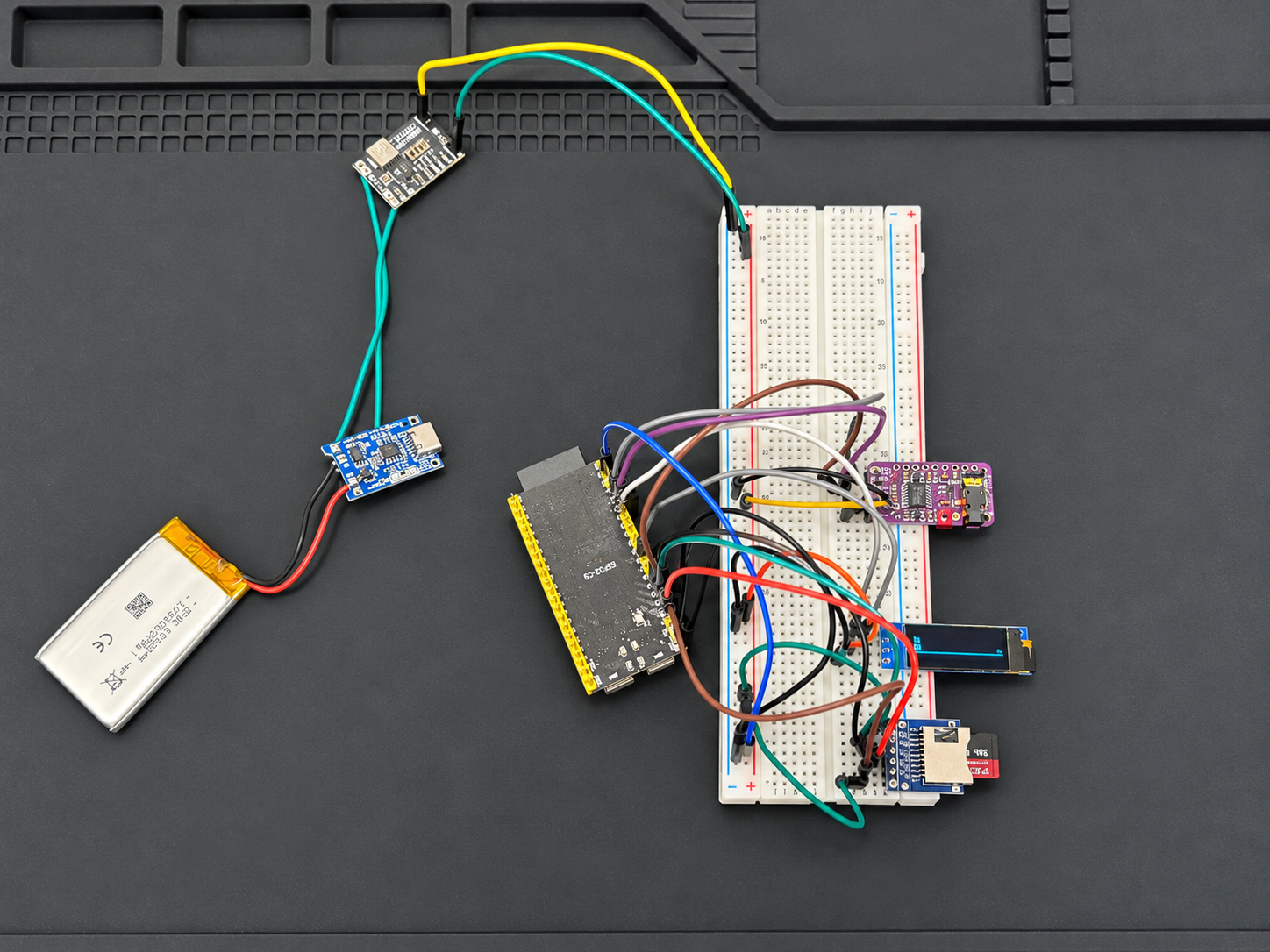 MP3 player prototype on a work mat, with a wired breadboard, microcontroller, small illuminated display, microSD module, battery, and connected boards.